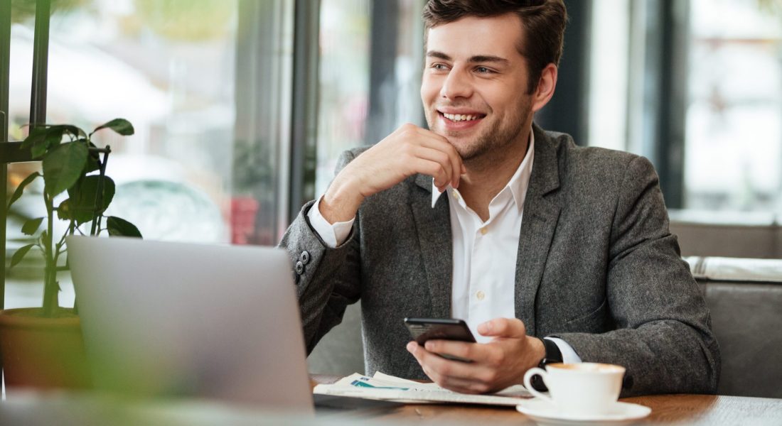 Smiling business man sitting by the table in cafe with laptop computer and smartphone while looking away